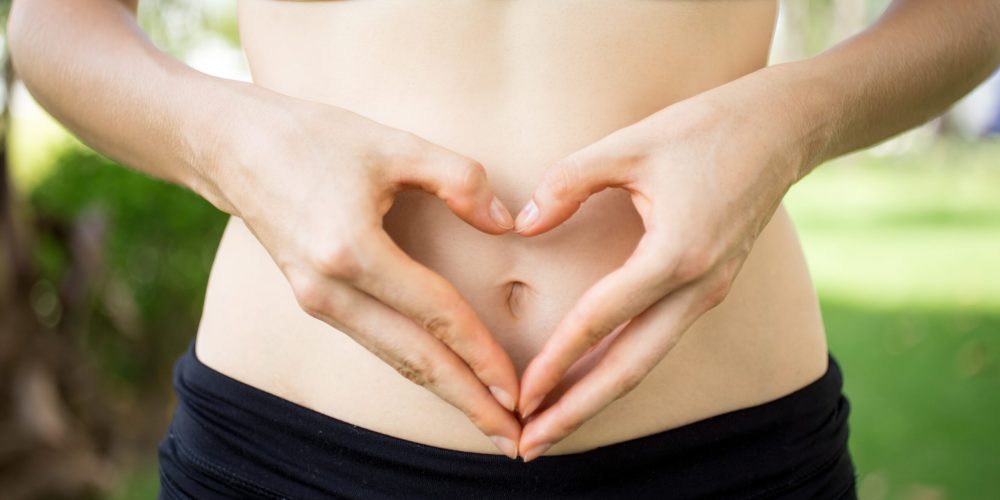 Close-up of female hands shaping heart on belly outdoors. Fitness and dieting concept